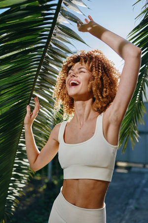 young woman with curly hair enjoys sunshine outdoors, wearing white sportswear near palm leaves,...