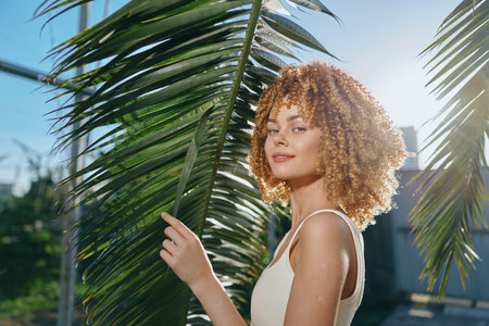 curly-haired woman in white top holding palm leaf outdoors, sunny day with blue sky, natural light enhancing her confident and relaxed expressionの写真素材