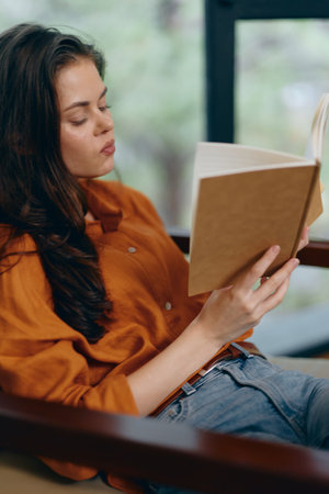 A young woman with long dark hair reading a book in a cozy setting, wearing an orange shirt and casual jeans, expressing calm and focus through her serene expressionの写真素材