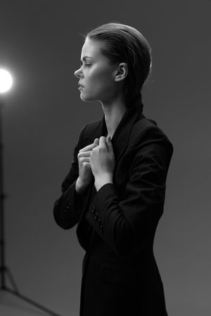 black and white serious woman dressed in black jacket standing sideways with hands near collar under soft studio light, portraying focus and calm moodの写真素材