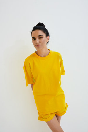 Young woman wearing bright yellow casual clothes stands against white background with relaxed expression, natural makeup, and tied-back hair in minimalistic studio setting.の写真素材