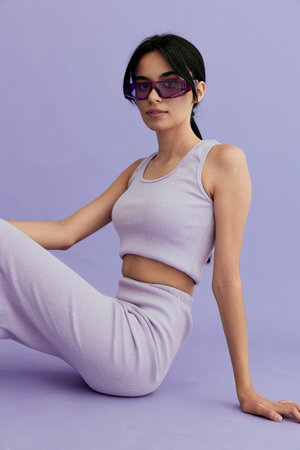 young woman sitting wearing lilac sleeveless top and matching pants with purple sunglasses against pastel purple background, casual relaxed pose and modern styleの写真素材
