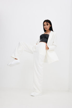 Young woman posing confidently in stylish white suit and black top with casual white sneakers against minimalist white background, showcasing modern fashion and elegance.の写真素材
