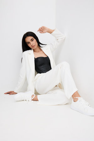 Young woman in white suit and black top sitting on floor against white wall, casual modern style with relaxed mood and minimalist bright background.の写真素材