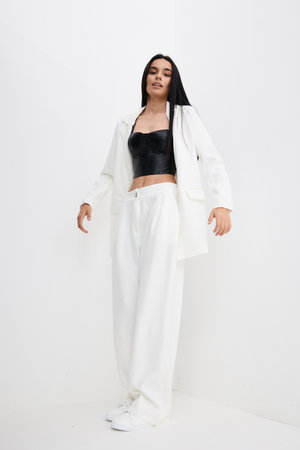Young woman in white oversized pants and blazer with black leather corset top posing confidently against minimalistic white background, showcasing modern fashion style.の写真素材