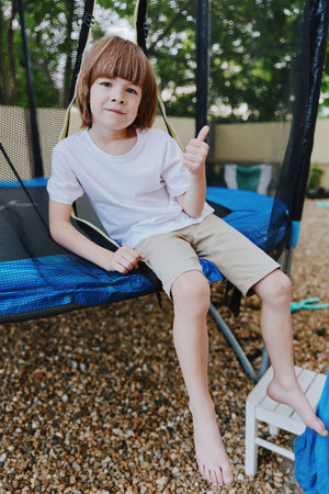 Cheerful boy giving a thumbs-up while sitting on a trampoline. The yard features gravel and greenery, creating an inviting outdoor atmosphere for play.の写真素材