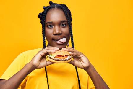 Young Black woman enjoying a colorful burger against a bright yellow background, showcasing joy and excitement with a playful expressionの写真素材