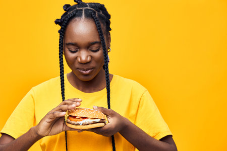 Young woman enjoying a delicious hamburger against a vibrant yellow background, showcasing healthy eating and happinessの写真素材