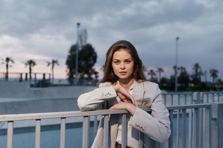 Woman in light suit leaning on metal railing outdoors with cloudy sky and palm trees in background, confident expression, business conceptの写真素材
