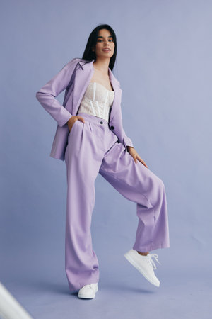 Young woman wearing a stylish lilac pantsuit and white sneakers posing confidently against a soft blue background. Modern, fashionable, poised, casual chic.の写真素材
