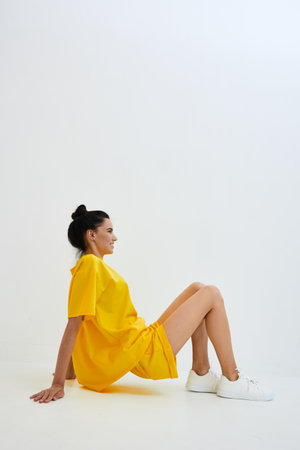 Young woman wearing bright yellow dress and white sneakers sitting on white floor in minimalistic studio, relaxed pose with casual style and soft lighting.の写真素材