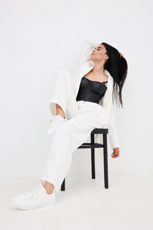 stylish woman in white pants and blazer poses seated on black chair with casual sneakers against plain white background, showcasing modern fashion and relaxed eleganceの写真素材
