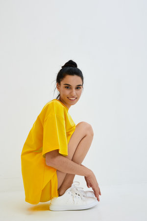 smiling woman wearing oversized yellow t-shirt and white sneakers squatting on white background with casual hairstyle, expressing relaxed and cheerful moodの写真素材