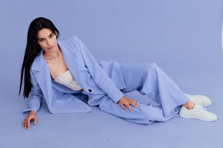 Young woman reclining on purple floor wearing pastel blue suit and white sneakers, dark hair,...