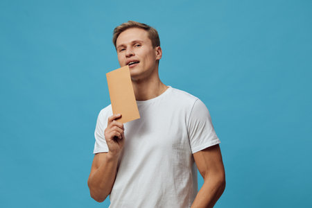 Young man holding blank cardboard sign in front of blue background, casual white t-shirt, smiling and looking at camera, advertising concept, studio shot, people lifestyle image, cheerful and friendly vibe.の写真素材