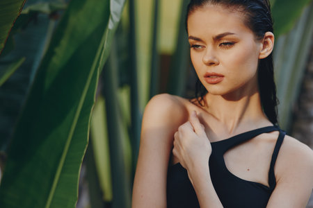 Attractive woman in black swimsuit posing confidently in front of lush palm tree in tropical paradiseの写真素材