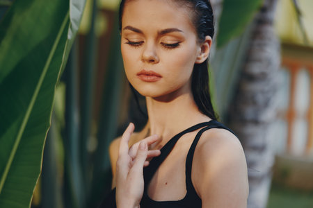 Beautiful woman in black tank top posing with hands on face in front of tall palm treeの写真素材