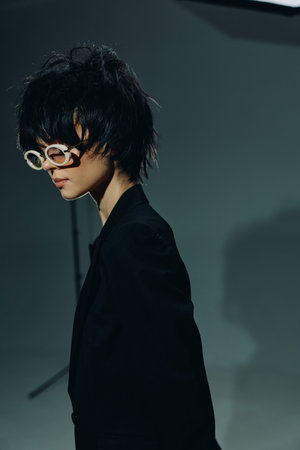 Young man wearing white round glasses and black suit jacket poses thoughtfully against a gray studio backdrop with soft lighting and moody atmosphere.の写真素材