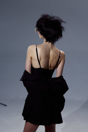 woman with short black hair wearing a black dress and glasses standing with her back against gray background in a studio settingの写真素材