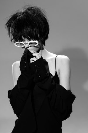 Black and white woman wearing white sunglasses and dark clothing posing with shy expression and short hair against plain background in stylish monochrome portrait.の写真素材