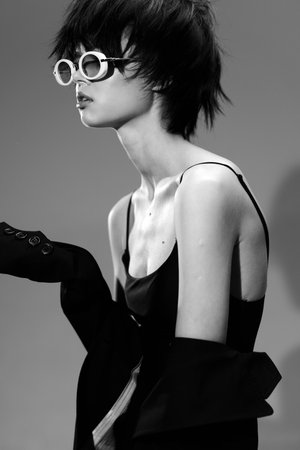 Black and white portrait of a young slender woman wearing round sunglasses and a black dress with off-shoulder sleeves, posing with an elegant and mysterious attitude.の写真素材