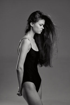 Black and white portrait of a slender woman in a black bodysuit with long flowing hair against a simple gray background, showcasing elegance and minimalistic style.の写真素材