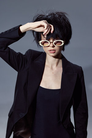 stylish woman wearing black blazer and white round sunglasses posing against gray background with cool confident expression and short dark hairの写真素材