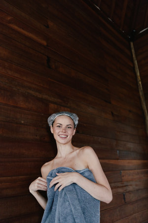 smiling woman wrapped in blue towel with hair net stands against dark wooden wall, natural light highlighting fresh skincare routine indoors, candid and relaxed.の写真素材