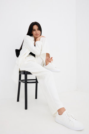 Young woman sitting on black chair wearing white suit and sneakers, posing in minimalistic bright studio, casual style and relaxed confident mood.の写真素材
