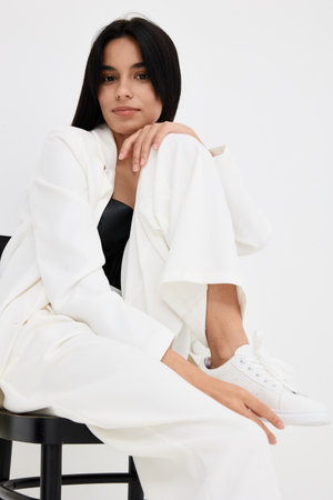 young woman in white suit and sneakers sitting on black stool with relaxed expression, modern casual fashion, minimalistic background, soft natural lightingの写真素材