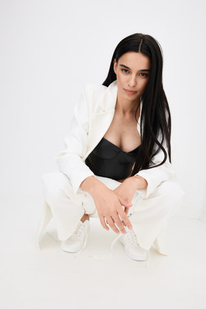 Stylish woman crouching in white suit and black corset top with long dark hair and clean minimalist background, conveying modern fashion and confidence.の写真素材