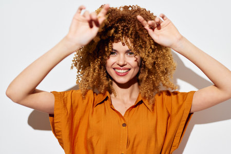 Smiling young woman with curly hair and an orange shirt posing playfully against a light background,...
