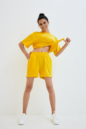 Smiling woman in bright yellow casual outfit posing confidently with tied shirt detail, white sneakers, and minimalistic white background, radiating energy and joy.の写真素材