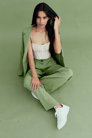 Stylish woman sitting on green floor wearing light jacket and pants with white sneakers, dark hair and confident pose in minimalistic studio setting.の写真素材