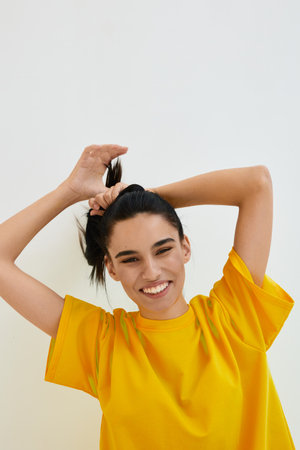 smiling woman in bright yellow t-shirt joyfully tying hair up against plain background, happy young adult expressing positivity and energyの写真素材