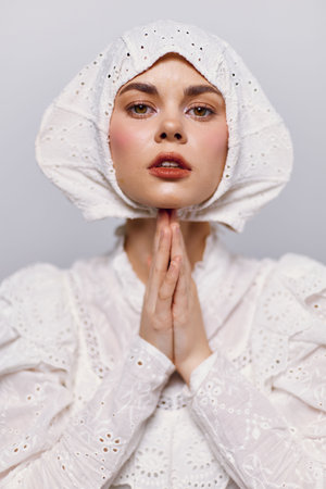 A young woman with a serene expression wears a vintage white outfit and a bonnet, showcasing a delicate and timeless style against a neutral backdropの写真素材