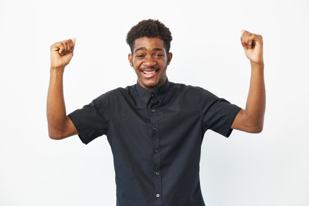 Happy African American man celebrating with raised arms, wearing a casual black shirt, exuding joy and enthusiasm in a bright atmosphere.の写真素材