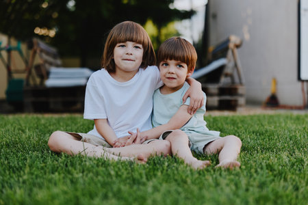 Two smiling children sitting together on the grass, enjoying a sunny day, showcasing friendship and joy in a vibrant outdoor setting.の写真素材
