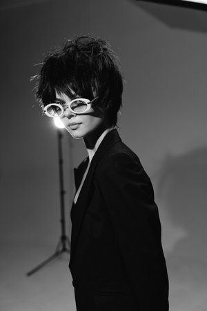 black and white woman wearing stylish sunglasses and dark blazer posing indoors with moody lighting and studio background in modern fashion portraitの写真素材