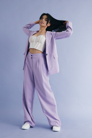 Stylish woman in lilac suit and white lace top posing confidently against soft lavender background, showcasing modern fashion and relaxed elegance in studio setting.の写真素材