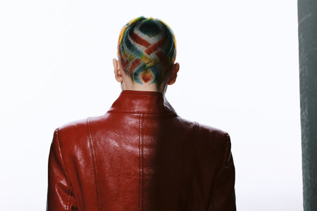 Woman with colorful geometric shaved hairstyle wearing a shiny red leather jacket standing against a...