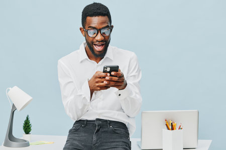 Smiling young man with glasses using smartphone in modern workspace. Casual business attire, bright minimalistic background, desk with laptop and stationery, cheerful mood. People lifestyle concept.の写真素材