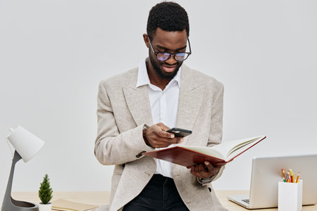 Professional young man using smartphone and reading a notebook while standing at a desk with a laptop and supplies, in a modern office environment. Business communication and technology concept, full body shot.の写真素材