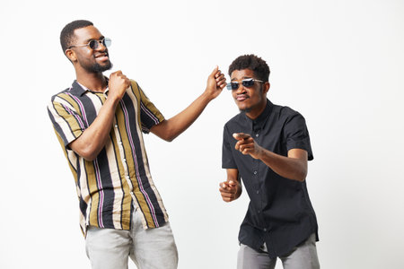 Two joyful black men dancing with sunglasses, showcasing friendship and fun. Their playful energy highlights modern fashion in a bright, minimalist setting.の写真素材