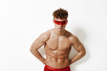 Confident young man with curly hair posing against a white background, wearing stylish red sunglasses and red swimwear. Fashion and self-expression concept.の写真素材
