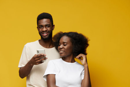 Two joyful young adults smiling together against a vibrant yellow background, showcasing modern casual attire and a playful relationshipの写真素材