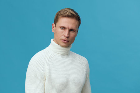 Close-up of serious young man with short blond hair wearing white turtleneck sweater against solid blue background, studio portrait, neutral expression, people lifestyle conceptの写真素材