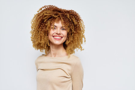 Happy woman with curly hair smiling against a light grey background, showcasing natural beauty and confidence in a casual light brown topの写真素材