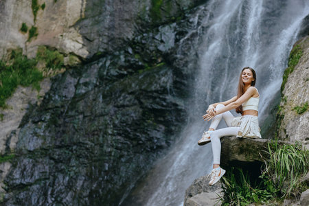 Smiling woman in casual white outfit sitting on rock near a waterfall, enjoying calm nature and refreshing outdoor atmosphere during a bright day.の写真素材