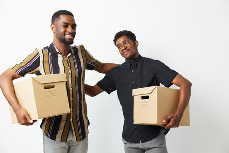 Two smiling African American men lifting cardboard boxes in a light and cheerful atmosphere, showcasing teamwork and friendship in a bright setting.の写真素材